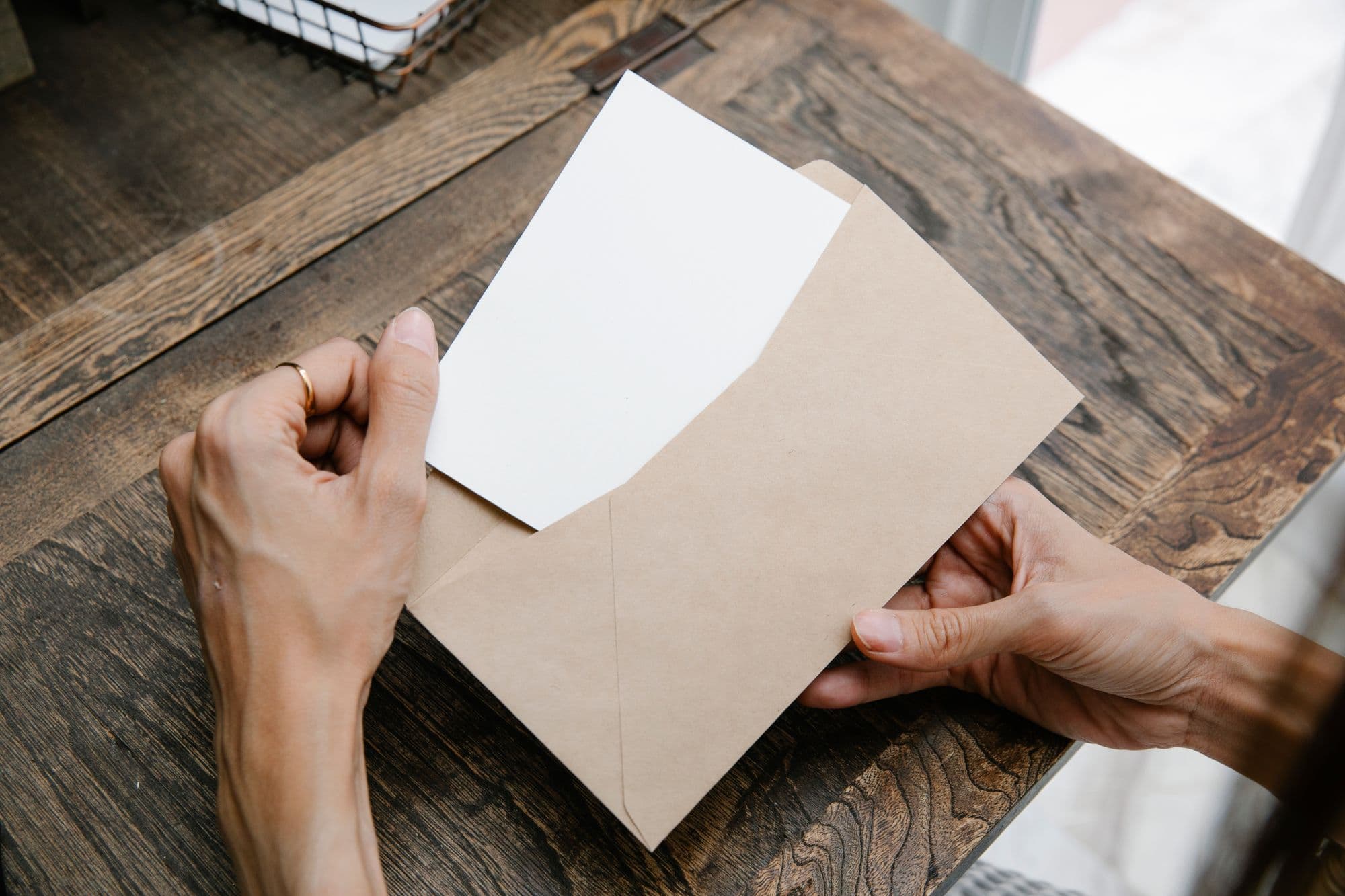 Des mains tiennent une lettre dans une enveloppe brune, vues d’en haut sur un bureau en bois.