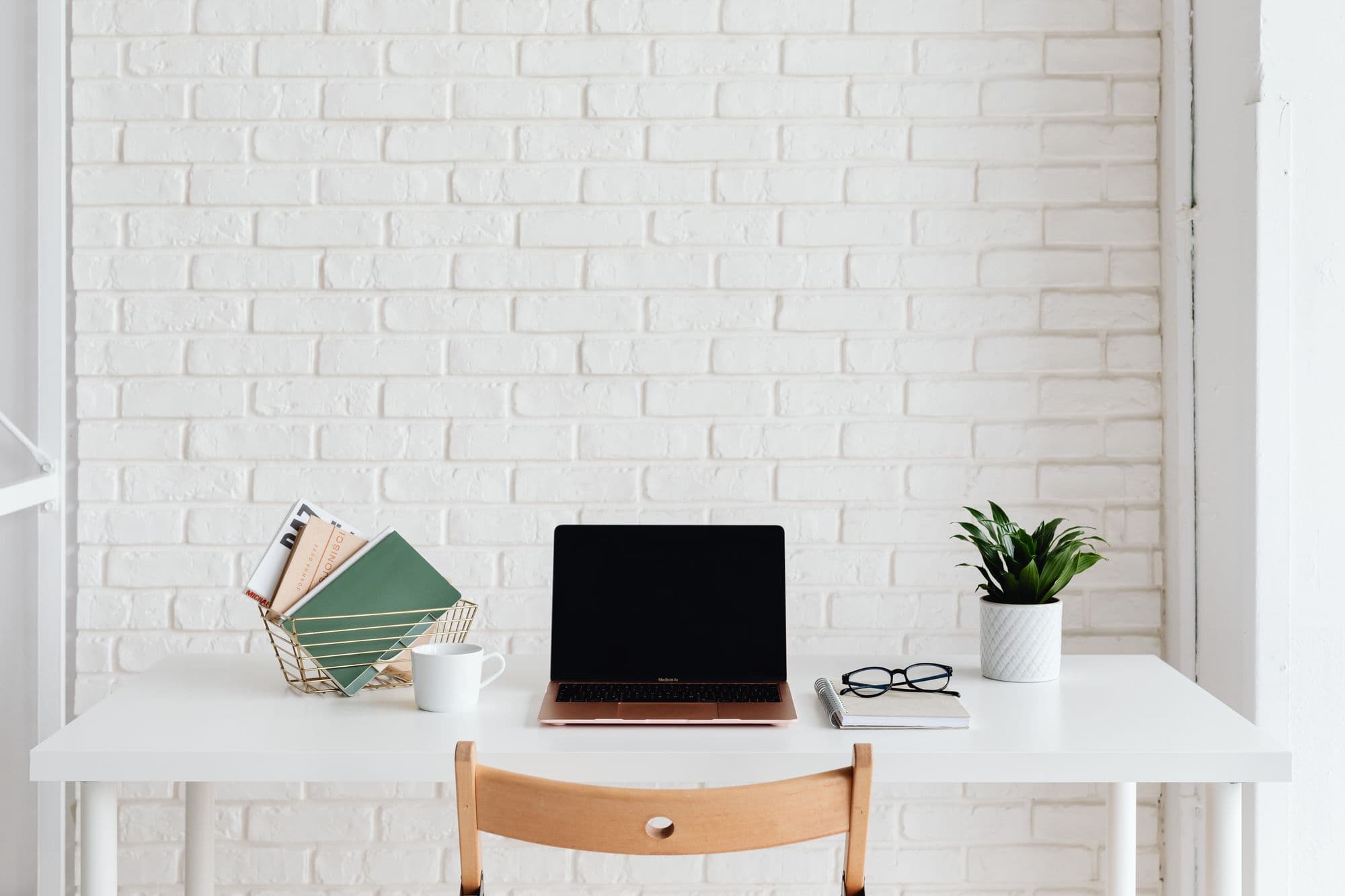 Un espace de travail blanc et ordonné, avec un ordinateur portable, une plante et des livres devant un mur de briques.